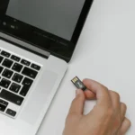Dłoń trzymająca niewielką pamięć USB obok otwartego laptopa