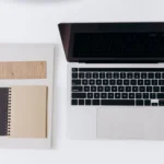 Laptop, notes w spirali i próbnik drewna ułożone na białym blacie, widok z góry